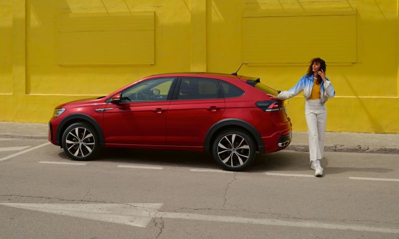 Un VW Taigo rouge au bord d’une route devant un immeuble jaune, vue latérale – une femme s’appuie sur l’arrière