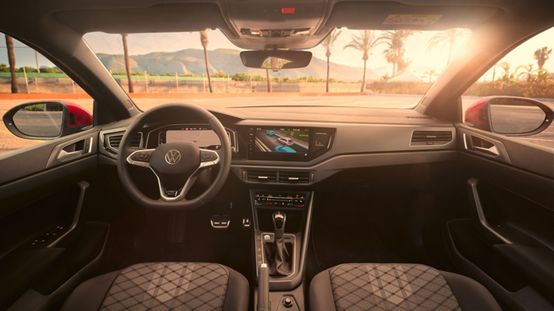 Blick auf das Cockpit mit Lenkrad und Infotainment-System im VW Taigo. Vor dem Fahrzeug sind Palmen und Berge.