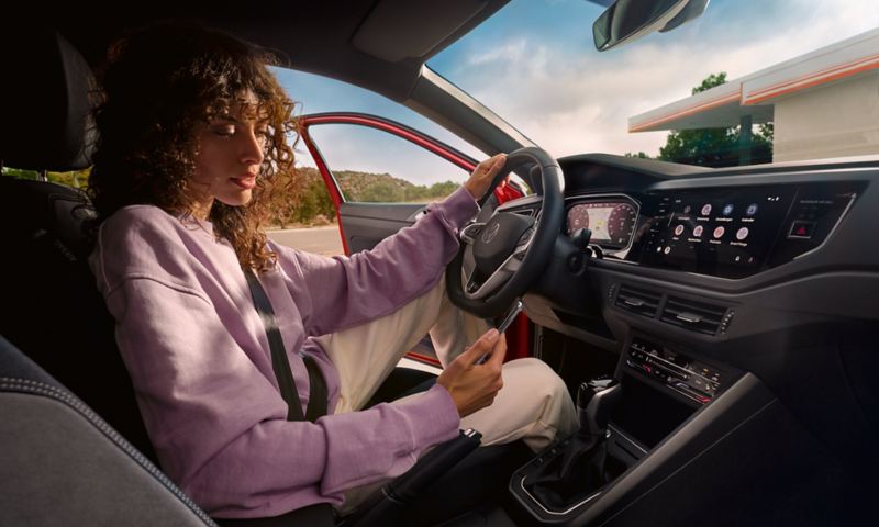 Interni della Volkswagen Taigo: una donna è seduta sul sedile del conducente di un’auto parcheggiata e guarda lo smartphone