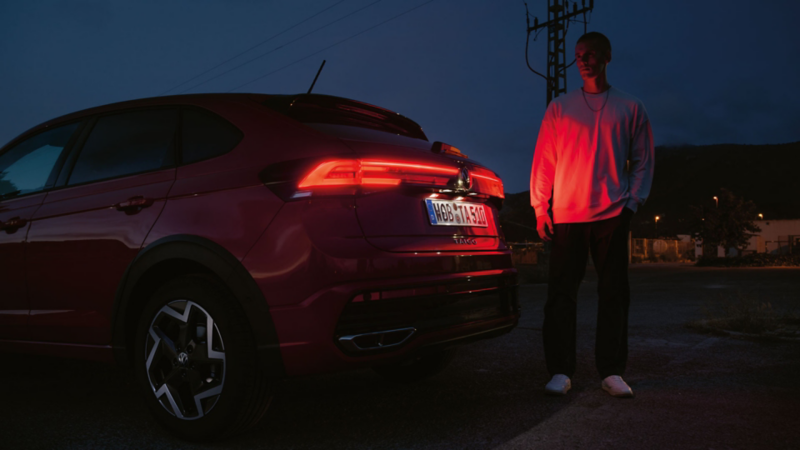 Ein Mann steht am Heck eines VW Taigo im Dunkeln und wird von den Rückleuchten rot angeleuchtet.