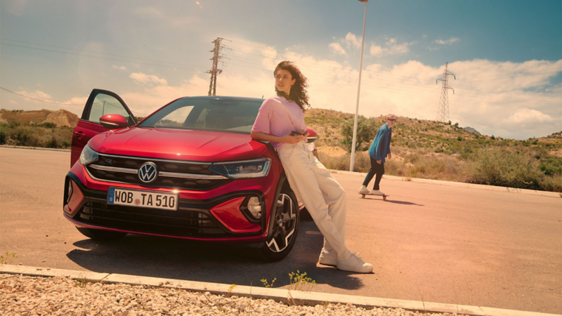 Frau lehnt an der Motorhaube eines VW Taigo auf einem Parkplatz in der Wüste. Mann fährt auf Skateboard dahinter entlang.