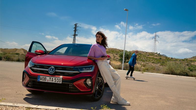 Rode VW Taigo R-Line, vooraanzicht op parkeerplaats. Vrouw leunt op motorkap, man rijdt op een skateboard