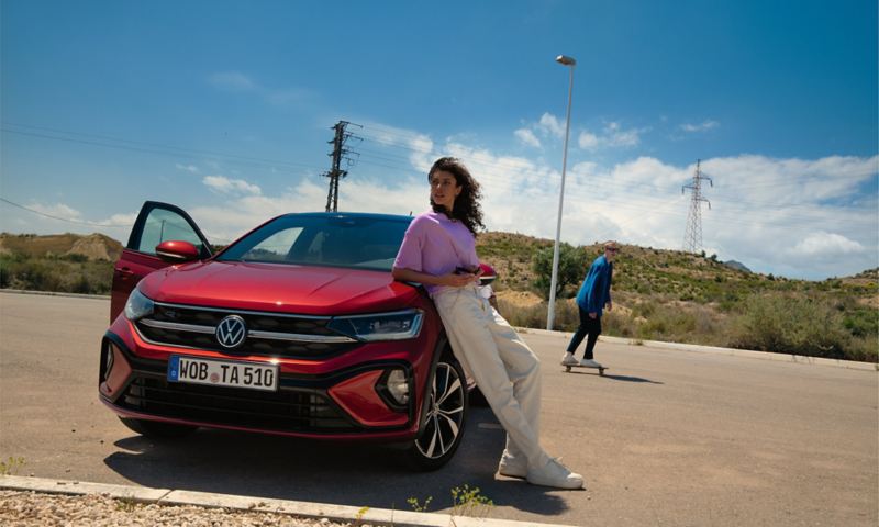 Front view of a red VW Taigo R-Line in a parking space. A woman is leaning on the bonnet, a man is skateboarding behind it