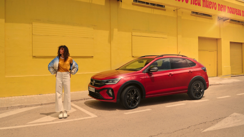 Picture shows Taigo R-Line in red with optional Black Style design package at the roadside in front of a yellow building, side view, a woman is standing next to the vehicle