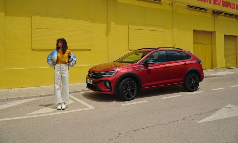 VW Taigo in Rot mit optionalem Design-Paket „Black Style“ am Straßenrand vor gelbem Gebäude, Seitenansicht, Frau steht neben Fahrzeug