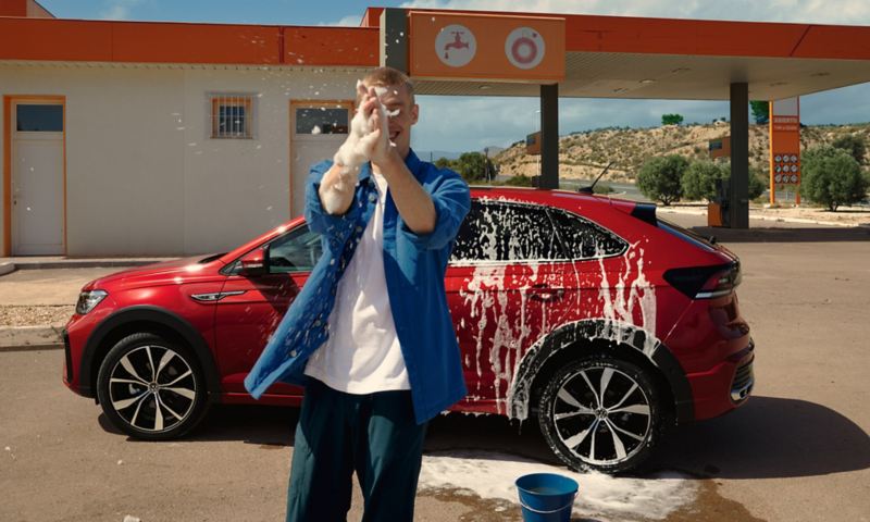Un VW Taigo rouge dans une installation de lavage de voitures, vue latérale, un homme joue avec de la mousse devant la voiture