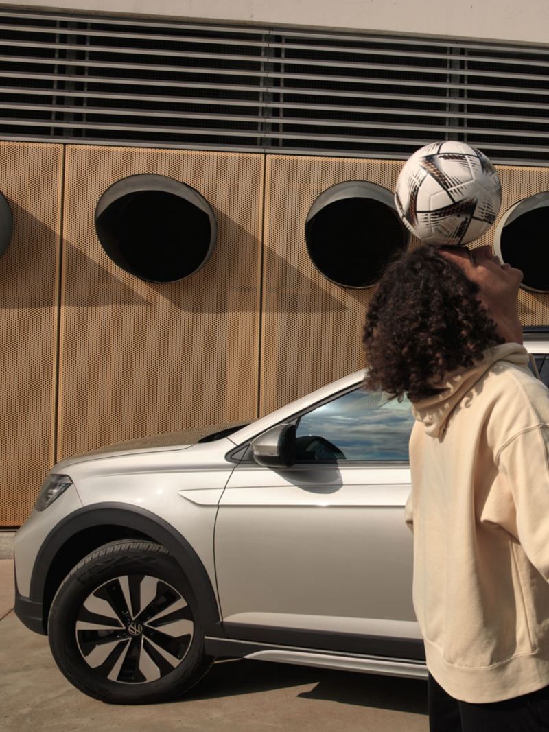 Ein Mann jongliert mit einem Fußball, im Hintergrund ein VW Taigo MOVE in silber, Blick auf Seite und Rückleuchte