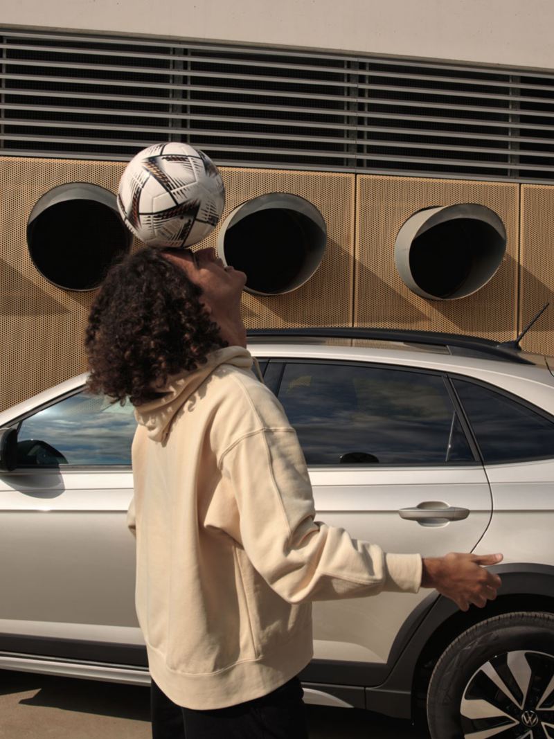 Ein Mann jongliert mit einem Fußball, im Hintergrund ein VW Taigo MOVE in silber, Blick auf Seite und Rückleuchte