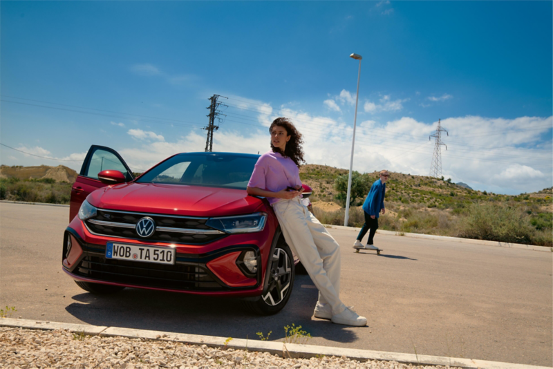 Vista frontale di una VW Taigo R-Line rossa in un parcheggio. Una donna è appoggiata al cofano, un uomo sta facendo skateboard dietro di lei