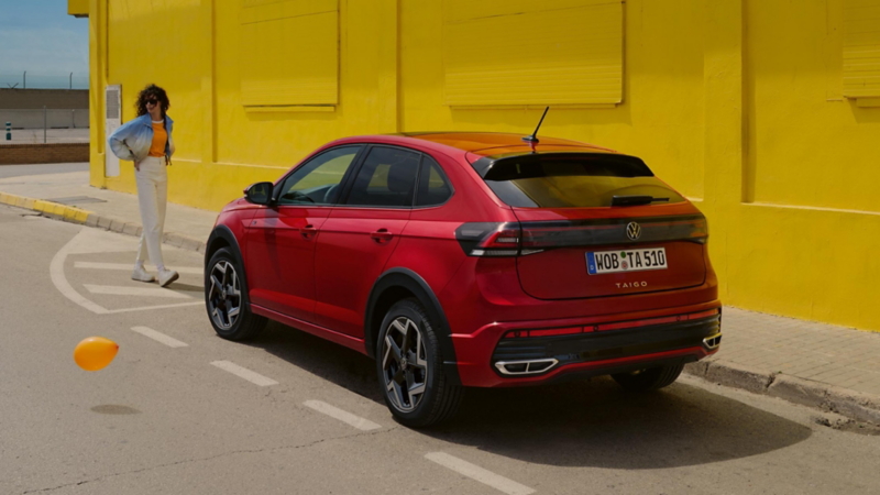 VW Taigo vista di 3/4 posteriore parcheggiata lungo la strada accanto ad una parete gialla. Una donna è in piedi vicino al ciglio della strada
