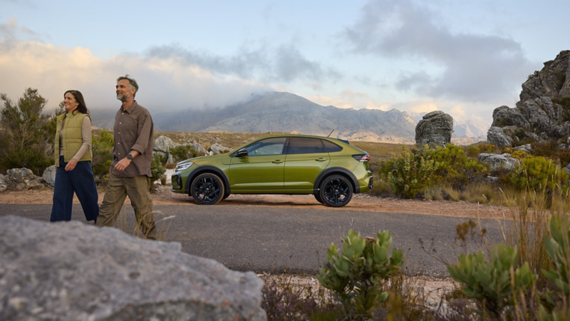 A couple walks past a parked green Volkswagen SUV in a rocky mountain landscape.