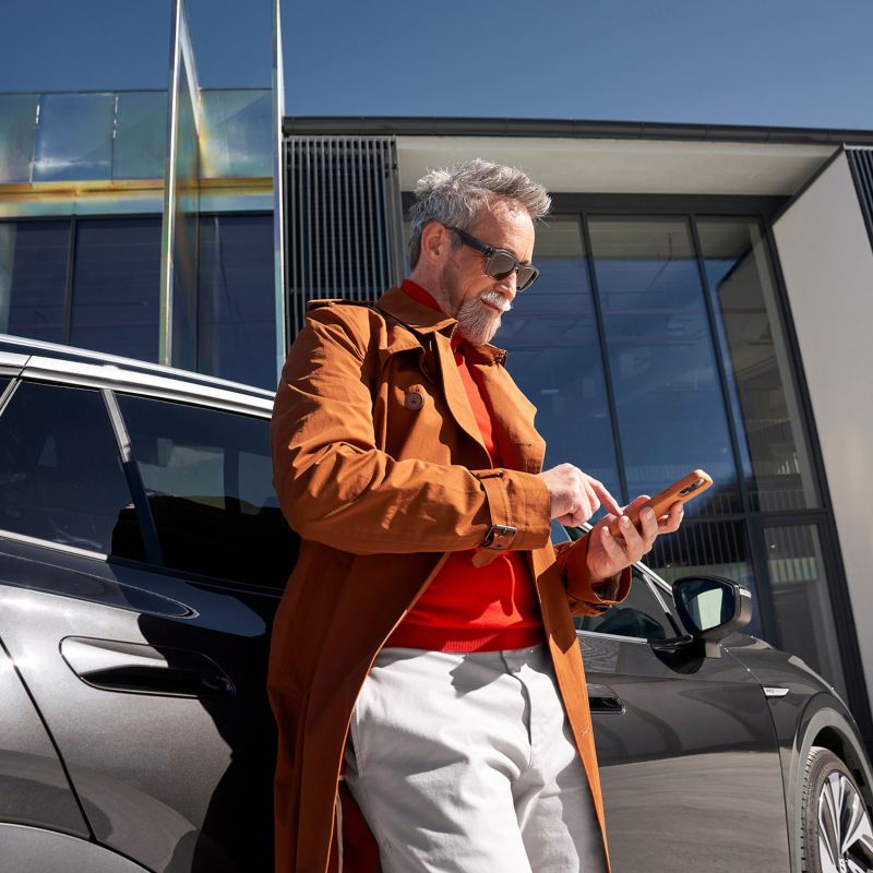 Ein Mann steht vor seinem VW ID.4 und schaut in sein Smartphone.