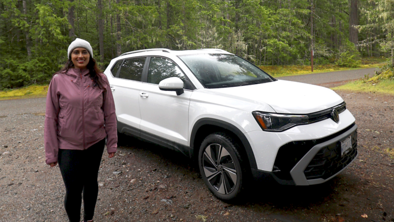 A woman in a rain jacket and toque smiles at the camera beside her white Atlas, parked on a dirt road in a forested area.