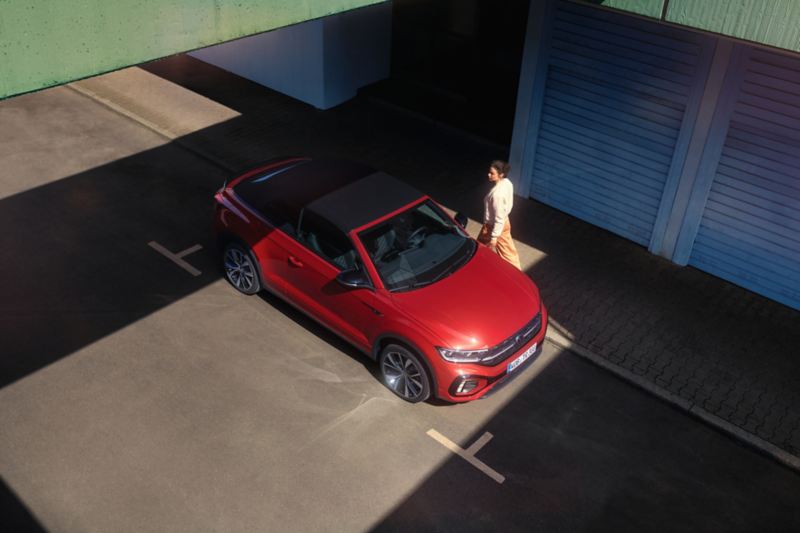 Bird's eye view of the red VW T-Roc Cabriolet with the roof open. Parking on the side of the street, a woman walks past.