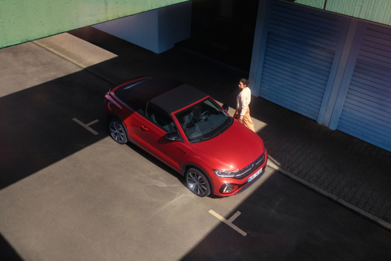 Vista aerea di VW T-Roc Cabriolet con il tetto aperto. Parcheggiata sul ciglio della strada, passa una donna.