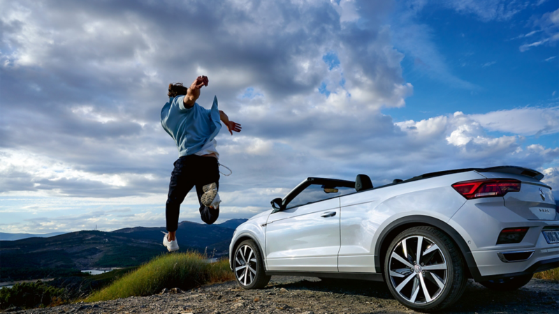 White Volkswagen convertible parked on a hill, with a man jumping, captured from behind