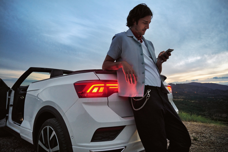Un homme en chemise décontractée est appuyé sur l'arrière d'une Volkswagen T-Roc Cabriolet blanche, regardant son téléphone, avec un paysage au crépuscule en arrière-plan.