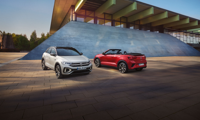 Ein weißer VW T-Roc mit schwarzem Dach und ein rotes VW T-Roc Cabriolet stehen vor einer Eventhalle.