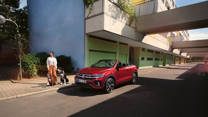 Rotes VW T-Roc Cabriolet mit geöffnetem Dach parkt am Straßenrand. Eine Frau mit zwei Hunden steht daneben.