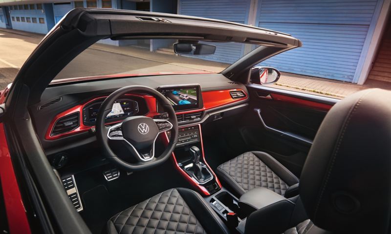 Interieur van de VW T-Roc Cabrio: zicht door het geopende dak van bovenuit op de cockpit met rood decor en voorstoelen.