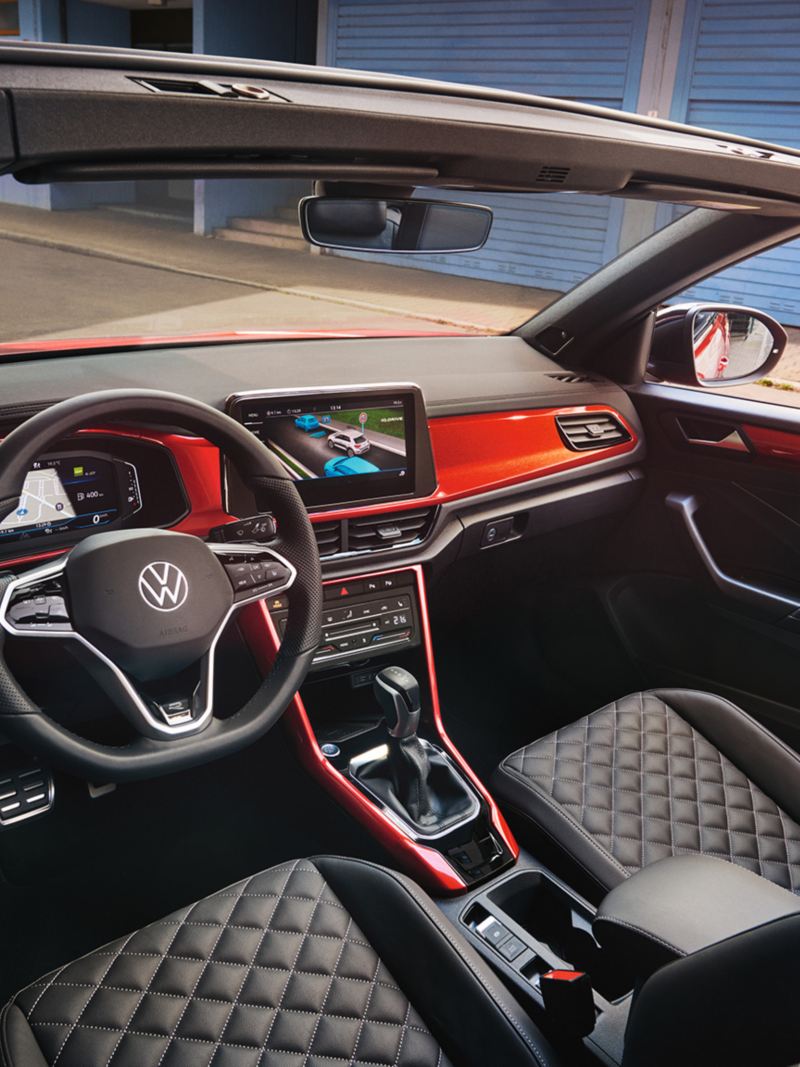 Vista degli interni, attraverso il tettuccio aperto, di Volkswagen Nuovo T-Roc Cabriolet, con dettaglio sul volante multifunzione e la plancia di comando.