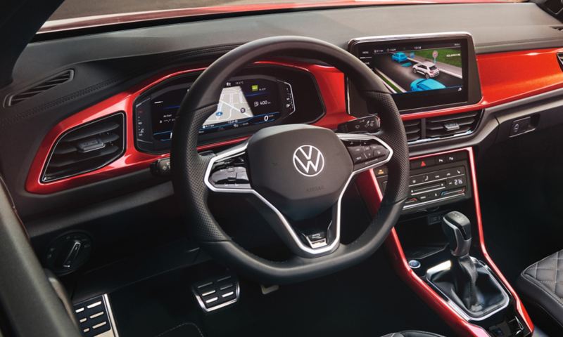 Interieur des VW T-Roc Cabriolet: Blick durch das geöffnete Dach von oben auf das Cockpit mit rotem Dekor und die vorderen Sitze. 
