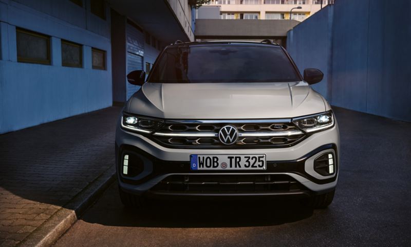 Front view of a VW T-Roc R-Line in grey, LED daytime running lights and the optional illuminated radiator grille are lighting