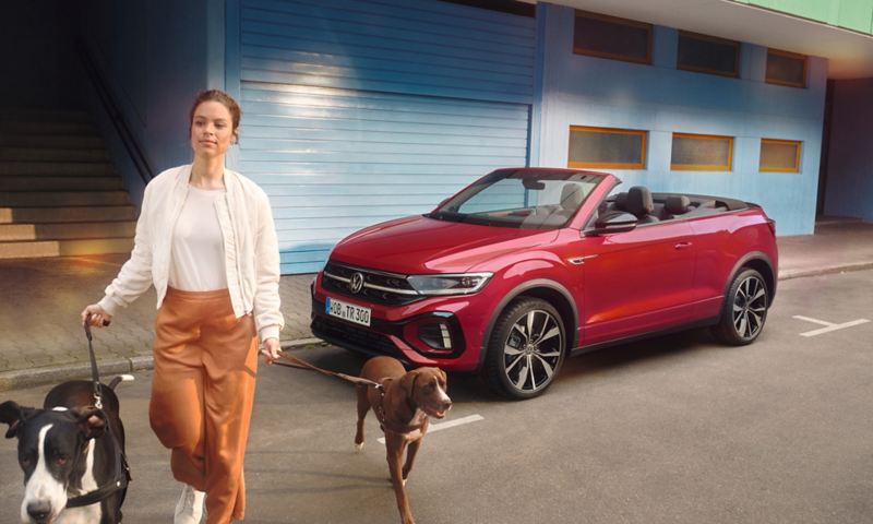 Red VW T-Roc Cabriolet with the roof open is parked on the side of the road. A woman with two dogs walks past it.