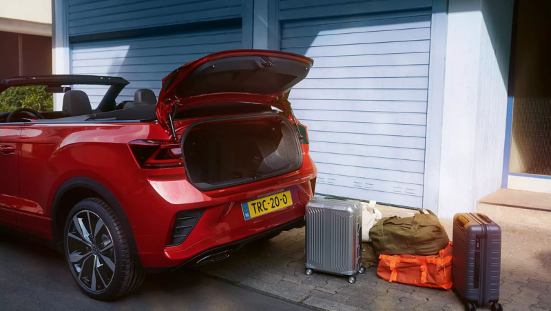 Schuin achteraanzicht van een rode Volkswagen T-Roc Cabrio R-Line met de kofferbak open, er staan verschillende koffers en tassen achter de auto