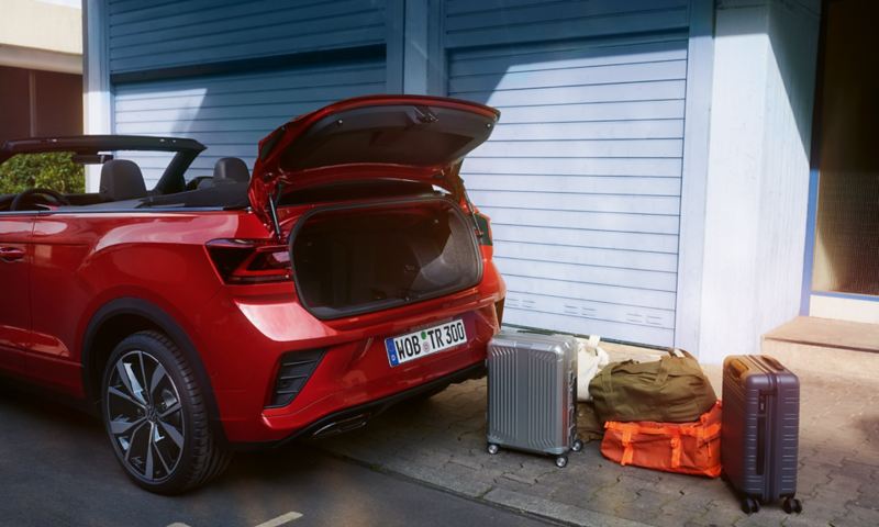 Volkswagen T-Roc Cabriolet rouge garé devant une sortie de garage avec le coffre ouvert et des bagages disposés derrière