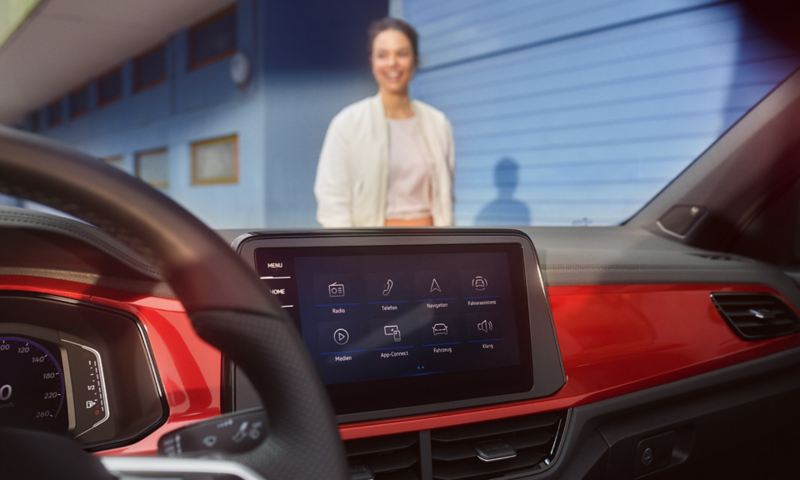 Intérieur du Volkswagen T-Roc cabriolet : Affichage du menu de système de radio & navigation et du volant multifonction. Une femme avance vers le véhicule.