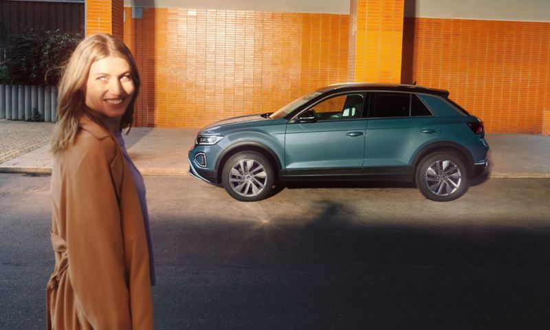 VW T-Roc Style бирюзового цвета перед зданием, вид сбоку, на переднем плане – улыбающаяся женщина, которая смотрит в объектив камеры