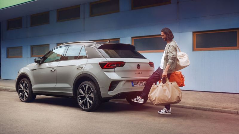 VW T-Roc R-Line in grey in front of a building, side visible, man standing behind the car and opening the boot lid with a swing of his foot