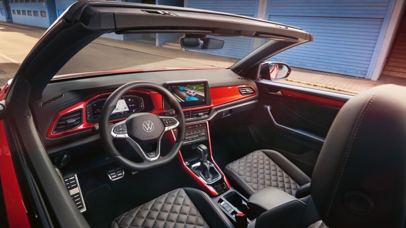 Interieur des VW T-Roc Cabriolet: Blick durch das geöffnete Dach von oben auf das Cockpit mit rotem Dekor und die vorderen Sitze. 