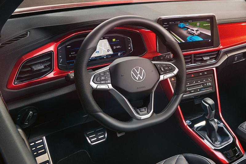 Interieur des VW T-Roc Cabriolet: Blick durch das geöffnete Dach von oben auf das Cockpit mit rotem Dekor und die vorderen Sitze. 