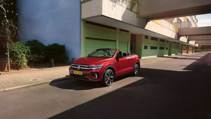Een rode VW T-Roc Cabriolet met het dak open staat langs de kant van de weg geparkeerd.