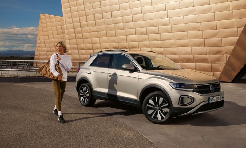 VW T-Roc MOVE in silber parkt vor einem Gebäude, Blick von der Seite auf die Frontscheinwerfer, eine junge Frau geht vorbei