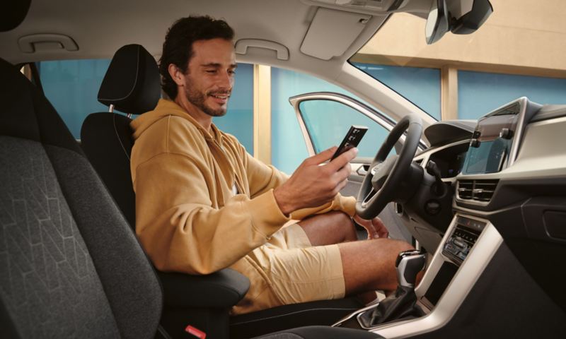 Blick in das Interieur des VW T-Roc Move, ein junger Mann in beiger Kleidung schaut bei geöffneter Tür auf sein Smartphone