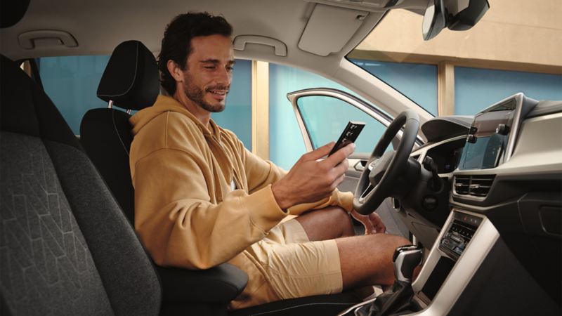 Vīrietis sēž Volkswagen automašīnā un skatās savā mobilajā tālrunī