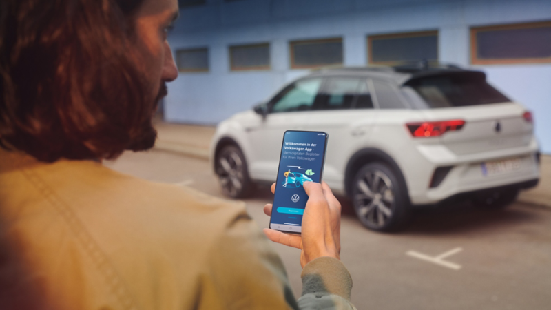 VW T-Roc R-Line in Grau vor einem Gebäude, Seite sichtbar, Mann steht mit Smartphone im Vordergrund.