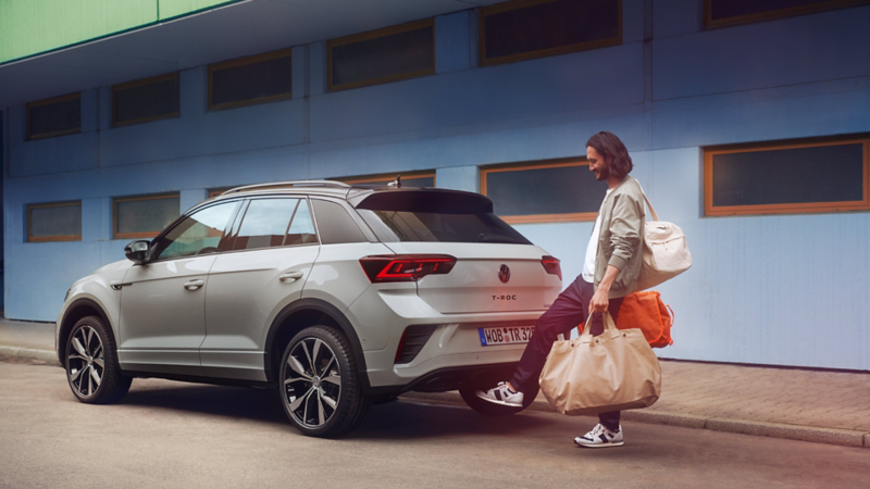 Ein grauer VW T-Roc R-Line parkt vor einem Gebäude. Eine Frau mit Taschen öffnet die Kofferraumklappe durch Fußschwenk.