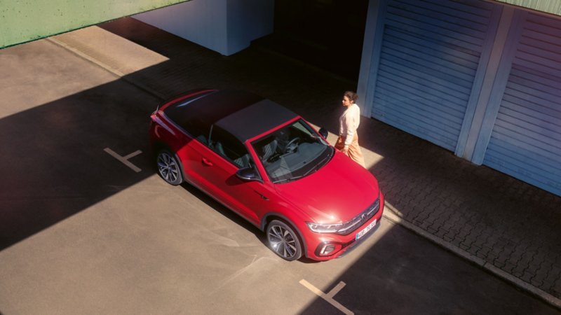 Vogelperspektive auf das rote VW T-Roc Cabriolet mit geöffnetem Dach. Parkend am Straßenrand, eine Frau geht vorbei. 