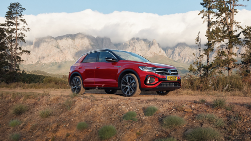 Un SUV Volkswagen T-Roc rouge est garé sur un chemin de terre, avec un arrière-plan composé de montagnes rocheuses et de nuages. La voiture est mise en valeur par la lumière naturelle et la couleur vive de sa carrosserie contraste avec la nature environnante.