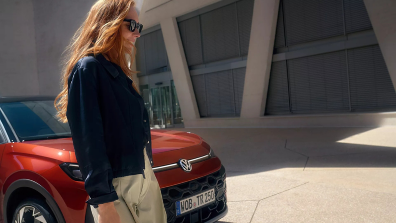 Kobieta w okularach przeciwsłonecznych stoi obok czerwonego VW T-Roc Style przed budynkiem.