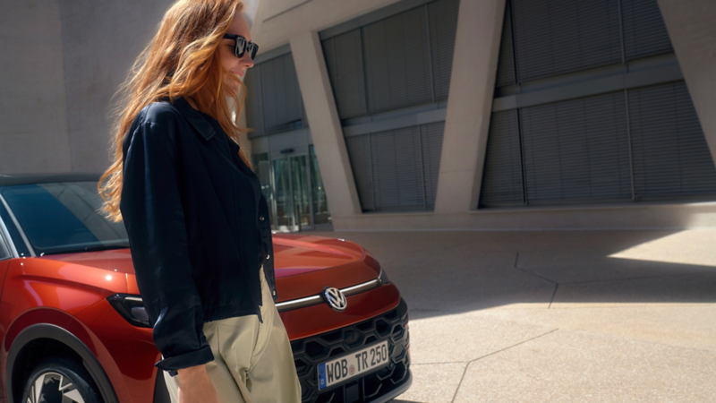 En kvinde med solbriller står ved siden af en rød Volkswagen T-Roc Style foran en bygning.