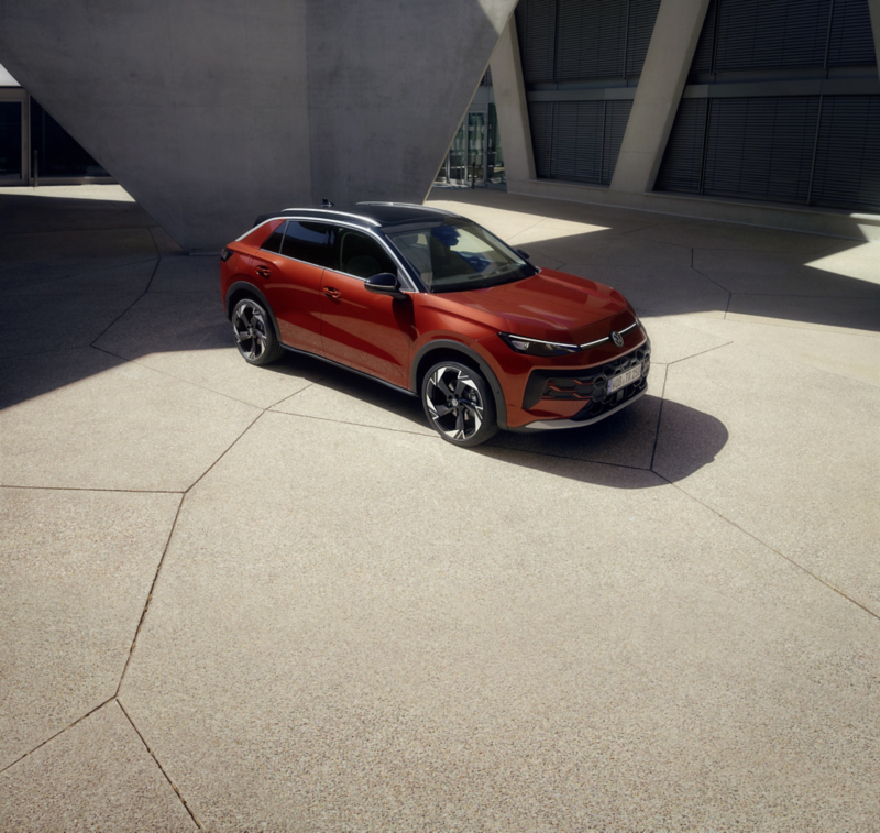 Un VW T-Roc Style de color rojo está aparcado frente a un edificio moderno bajo la luz del sol.