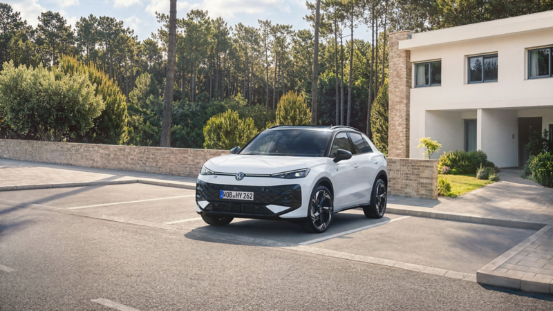 A white VW T-Roc hybrid is parked in a lot in front of a residential house by the forest, front view.