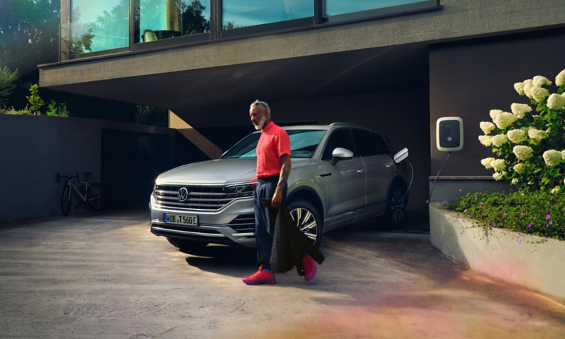 Volkswagen Touareg eHybrid in Silber, Sicht vorne-seitlich, lädt in Einfahrt, Mann geht vorbei
