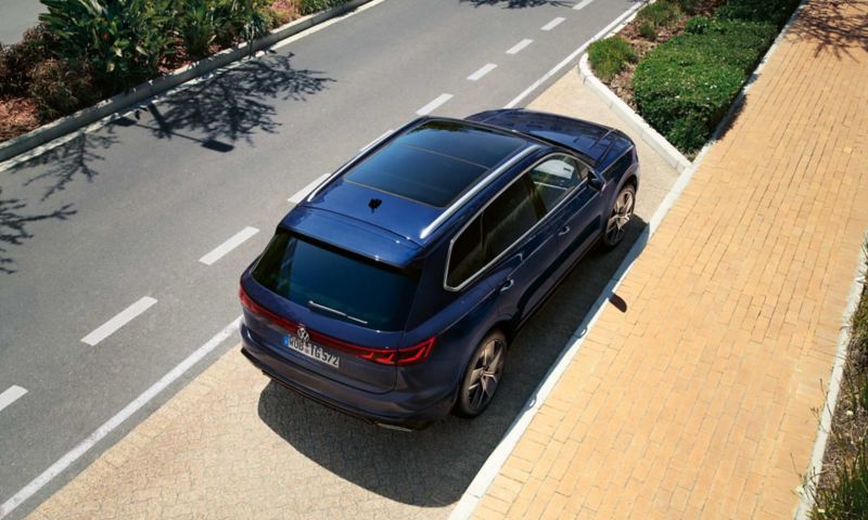 A VW Touareg R-Line parked at the roadside, diagonal view from above of the rear and panoramic sunroof.