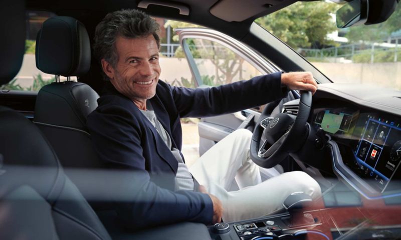Een man op de voorstoel van een geparkeerde VW Touareg R-Line kijkt glimlachend in de camera.